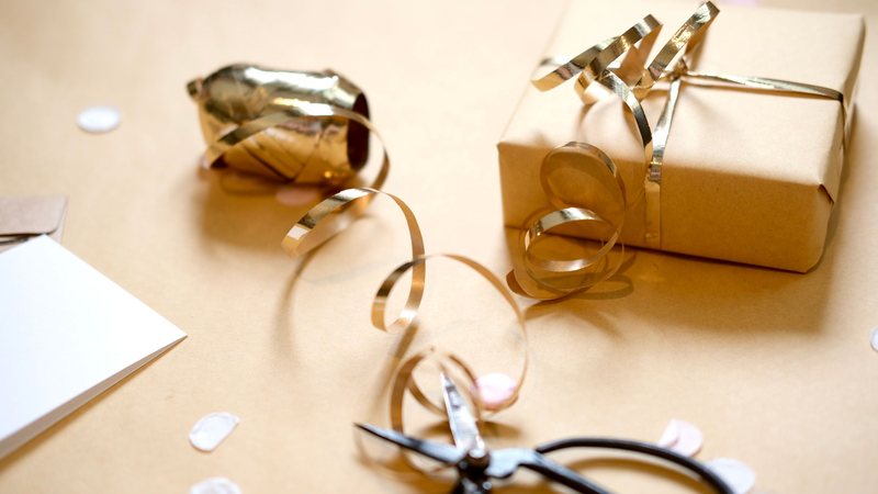 Photograph of a kraft-paper gift tied with curled gold ribbon next to a spool of gold ribbon, black scissors, a blank card and pale confetti