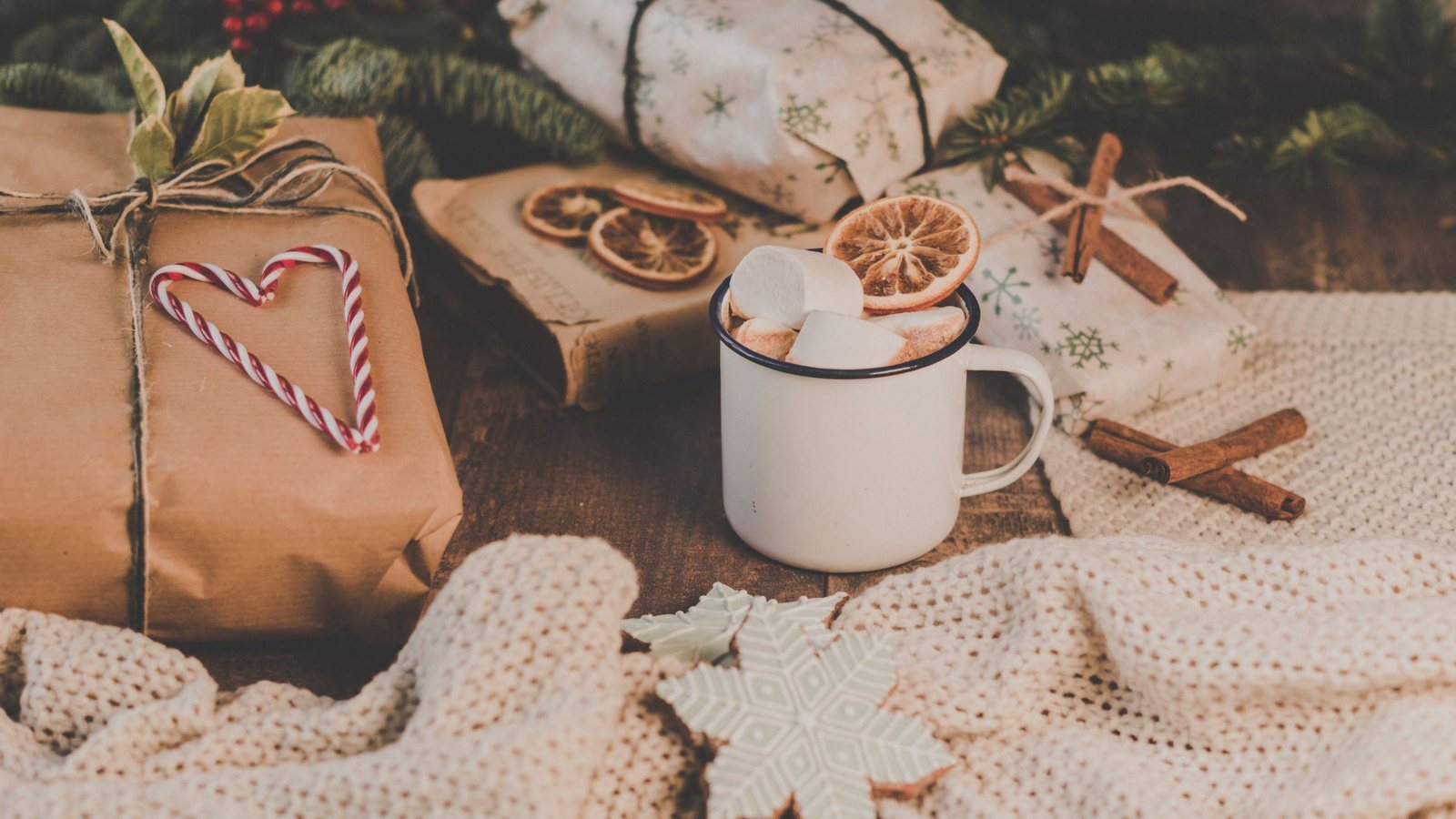 Photograph of an enamel mug of cocoa with marshmallows beside kraft-paper and printed-paper gifts tied with twine, cinnamon sticks and a cream knit throw