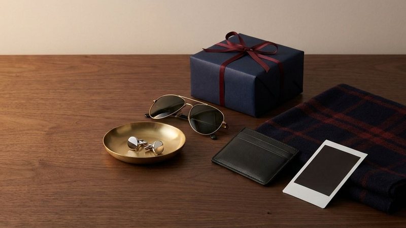 Still life of a navy wrapped gift, sunglasses, a brass dish, a leather card wallet and a plaid scarf on a dark wood table