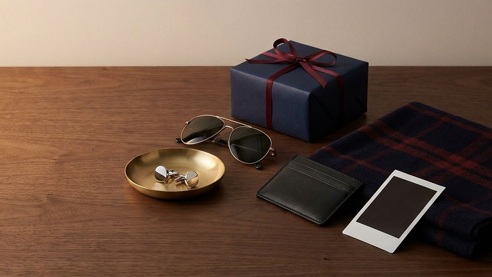 Still life of a navy wrapped gift, sunglasses, a brass dish, a leather card wallet and a plaid scarf on a dark wood table