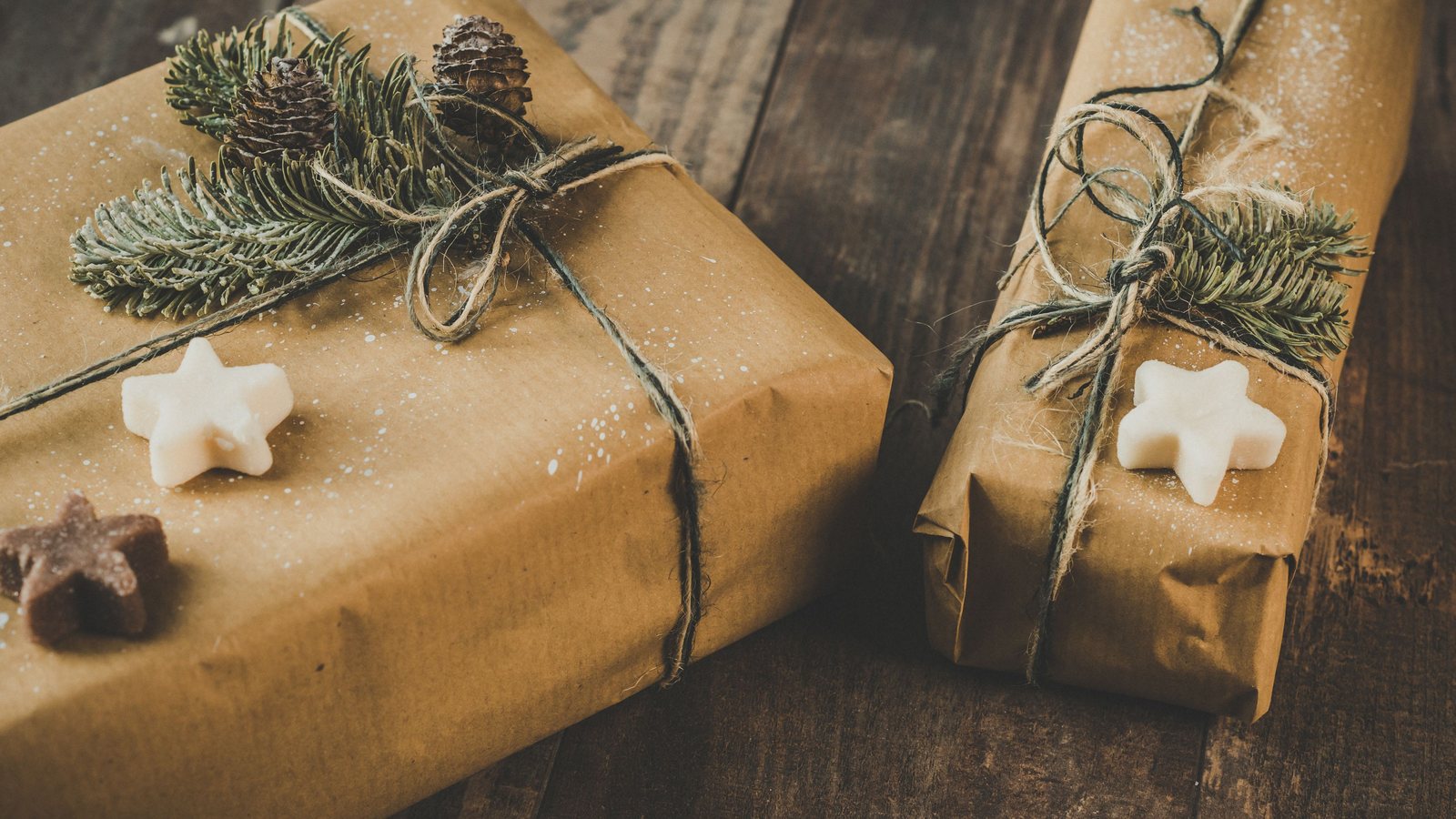 Photograph of two gifts wrapped in brown kraft paper and twine, topped with pine sprigs, pine cones and star-shaped cookies, on dark weathered wood