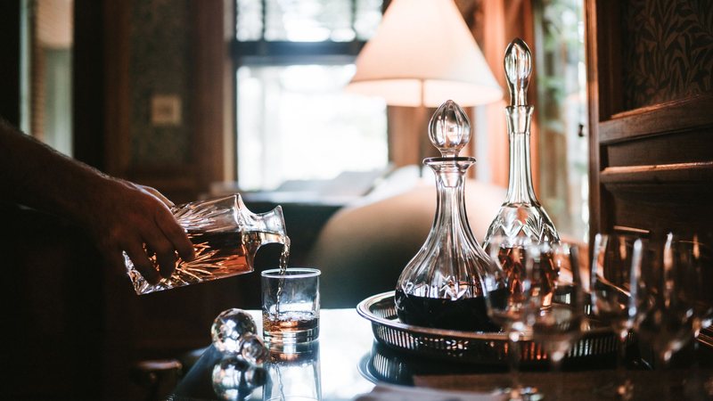 Photograph of a hand pouring whiskey from a crystal decanter into a tumbler in a lamplit room