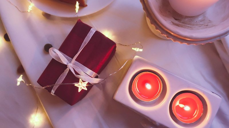 Photograph from above of a small gift wrapped in deep red paper and white ribbon beside two lit tealights and a string of star-shaped fairy lights on white linen