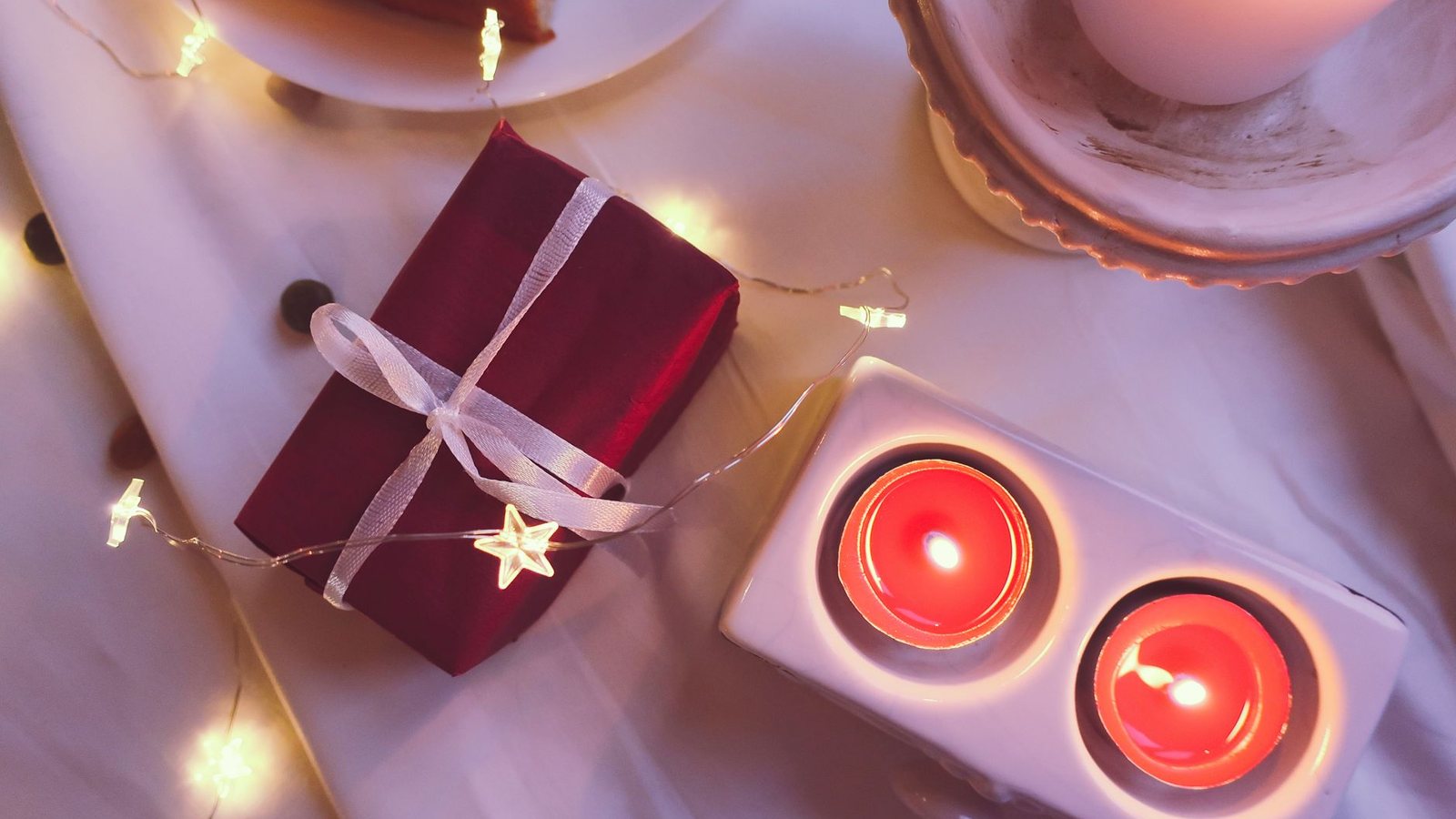 Photograph from above of a small gift wrapped in deep red paper and white ribbon beside two lit tealights and a string of star-shaped fairy lights on white linen