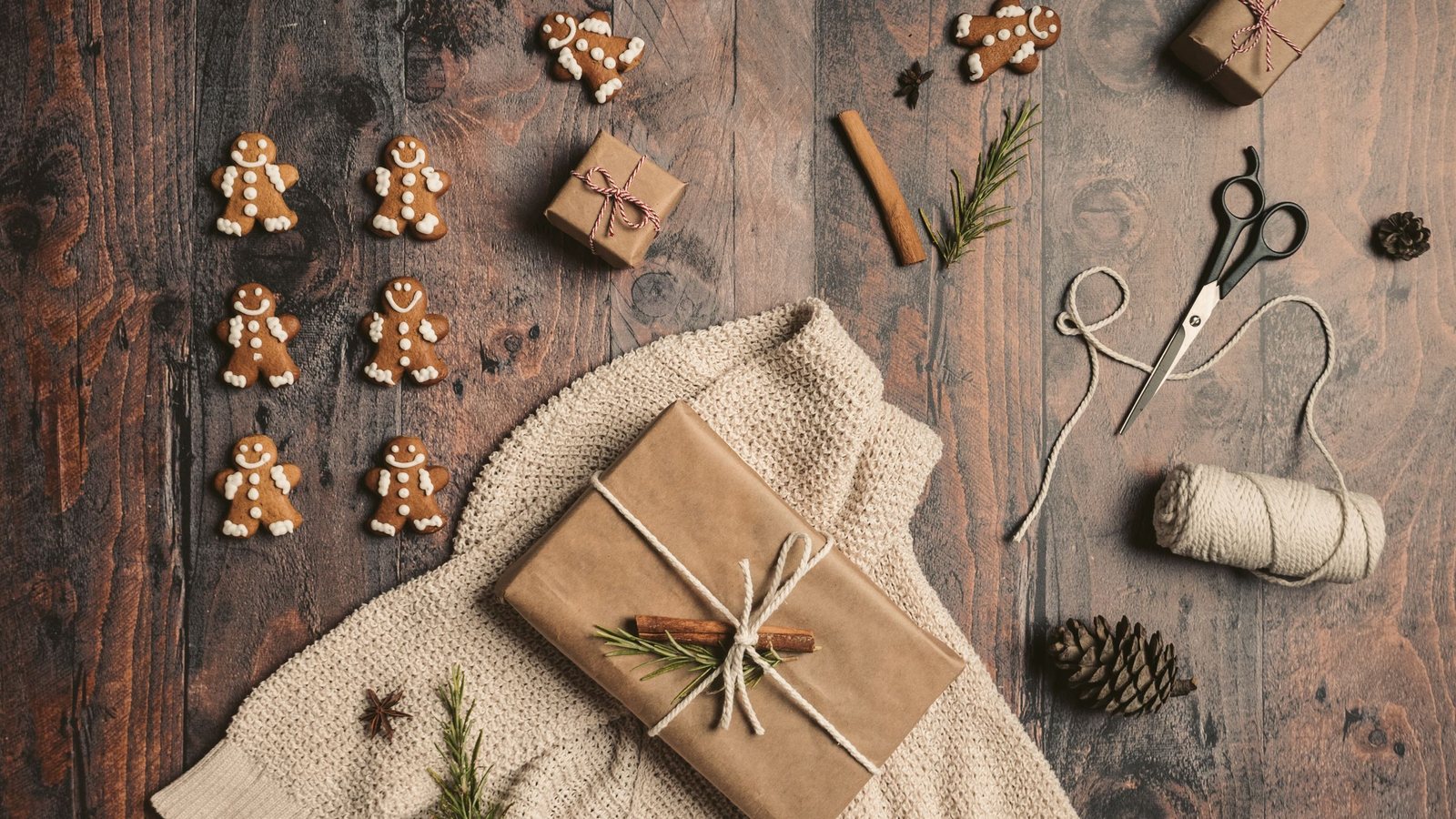 Photograph from above of a kraft-paper gift tied with twine on an oatmeal knit sweater, beside iced gingerbread cookies, scissors and a spool of twine on dark wood