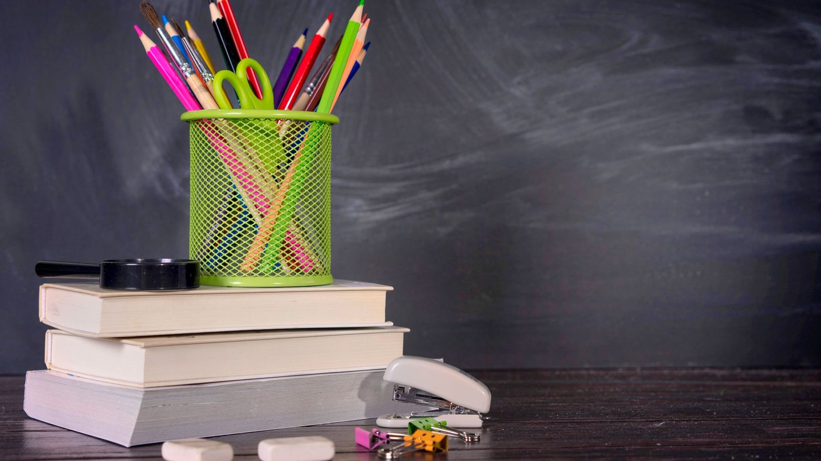 Photograph of colored pencils in a mesh cup on a stack of books in front of a chalkboard