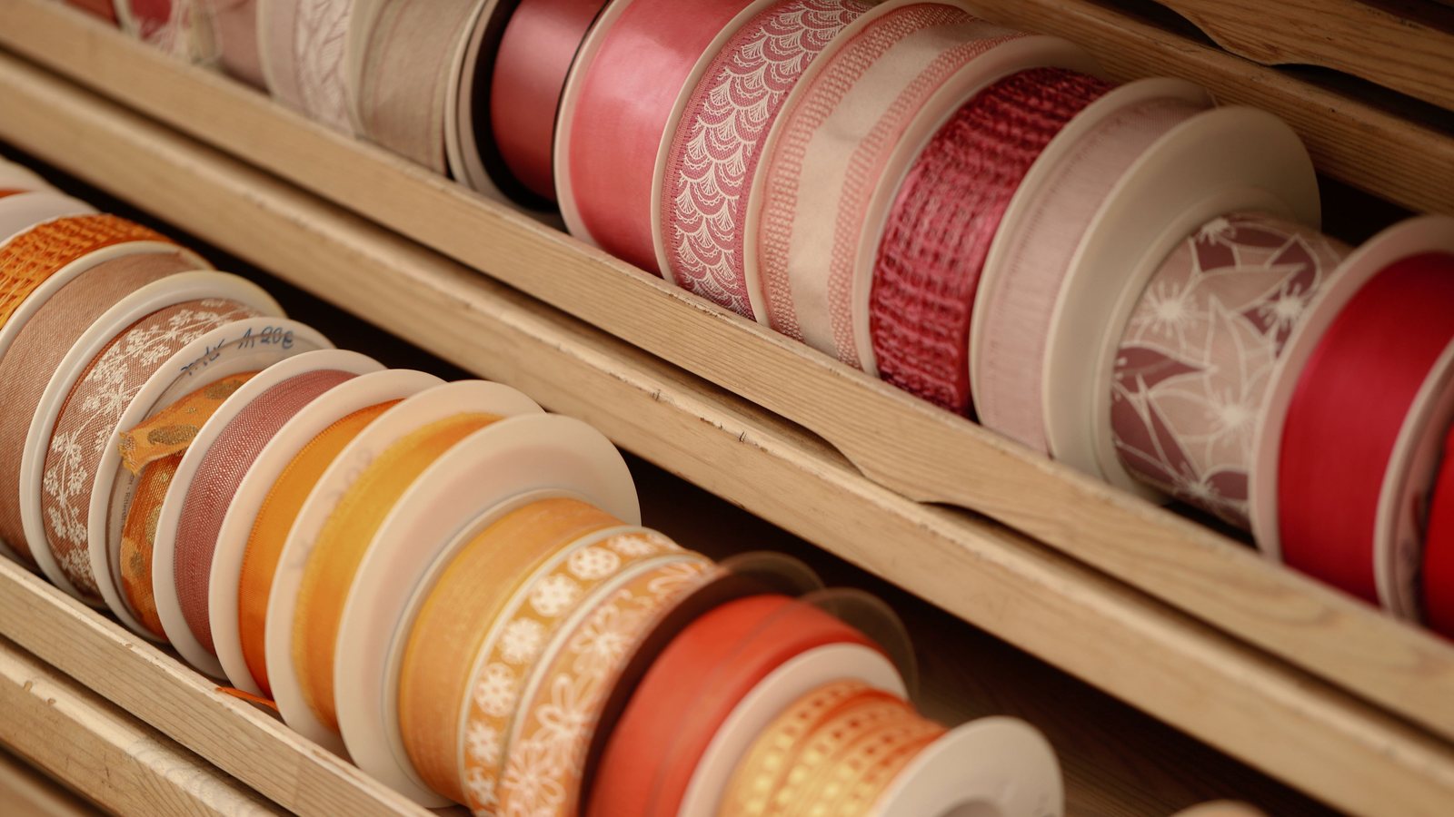 Photograph of rows of coral, deep red and cream ribbon spools stacked in a wooden rack