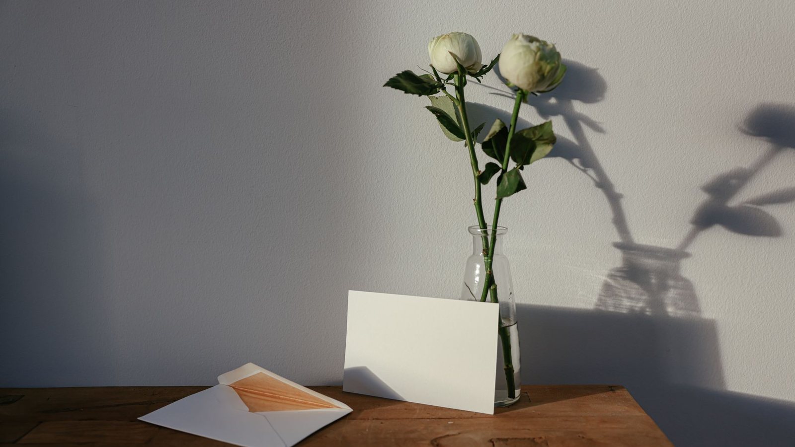 Photograph of a blank card propped against a glass bottle holding two white roses, beside an open envelope on a wooden bench in late afternoon sun