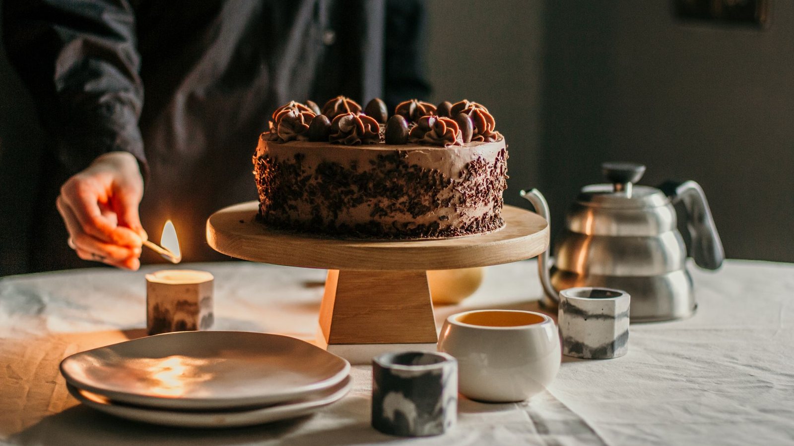 Photograph of a hand lighting a candle with a match beside a chocolate cake on a wooden cake stand, with cups and a steel kettle on a linen tablecloth