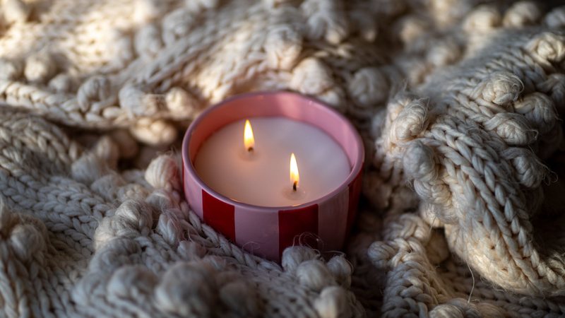 Photograph of a lit two-wick candle nestled in a chunky cream knit blanket