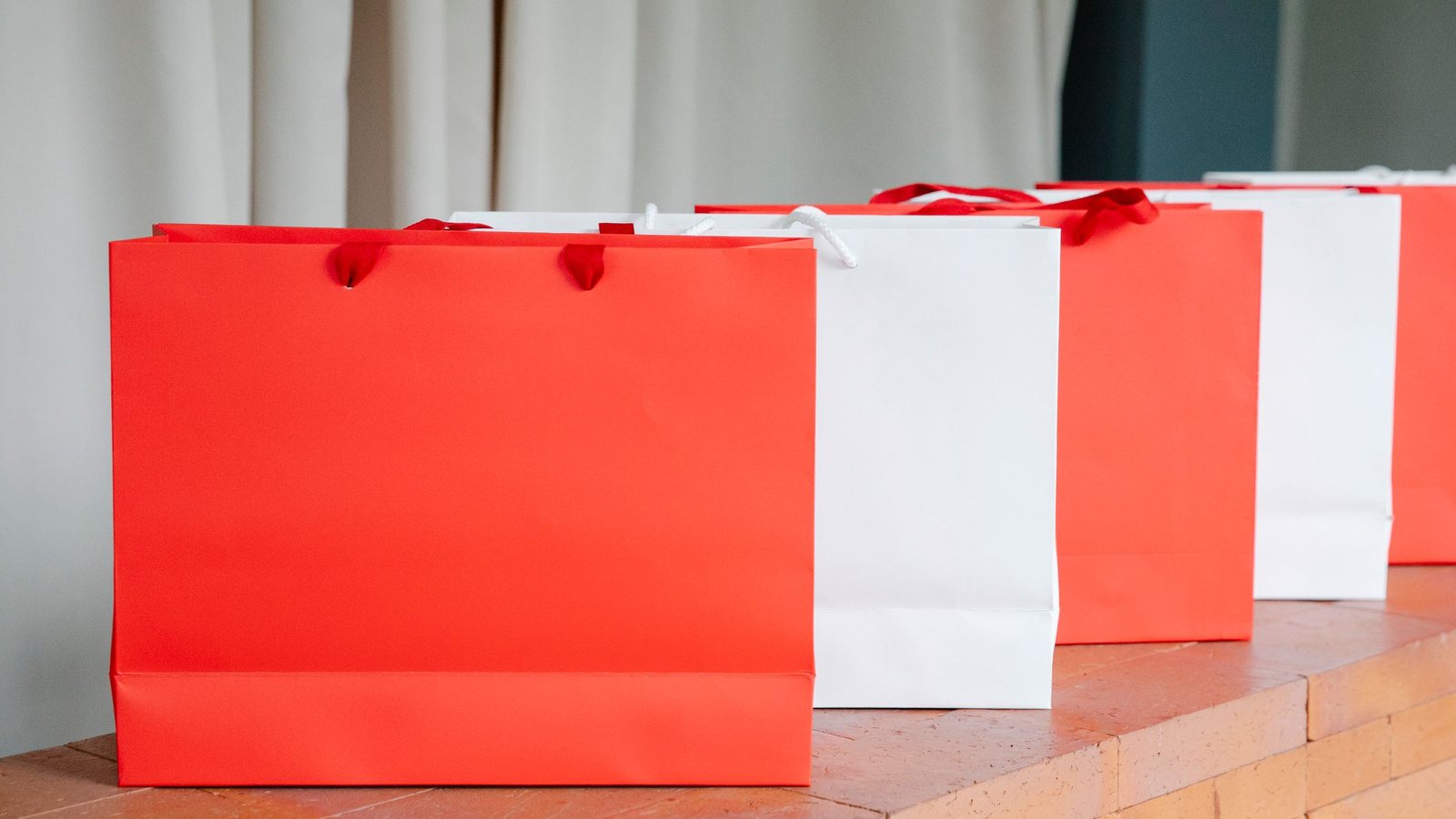 Photograph of red and white paper shopping bags lined up on a brick ledge in front of a white curtain