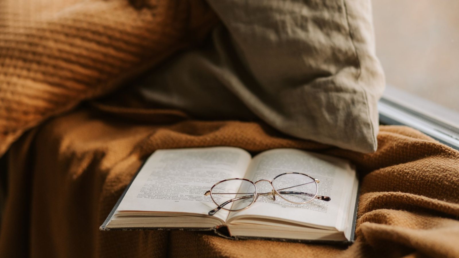 Photograph of an open book with reading glasses resting on a mustard knit throw beside a linen pillow