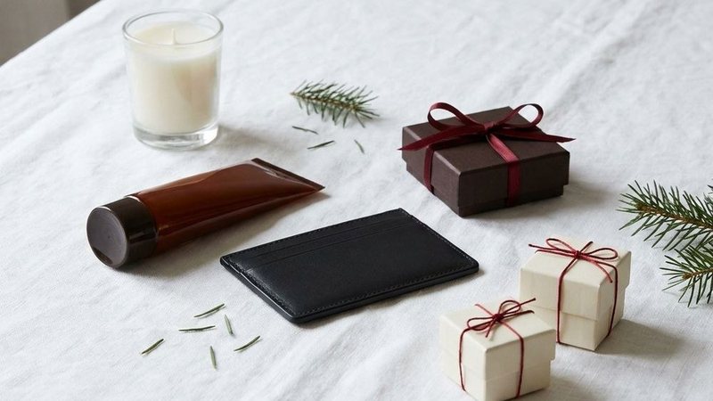 Still life of small ribbon-tied gift boxes, a candle, a leather card case and a grooming tube with fir sprigs on a white linen table