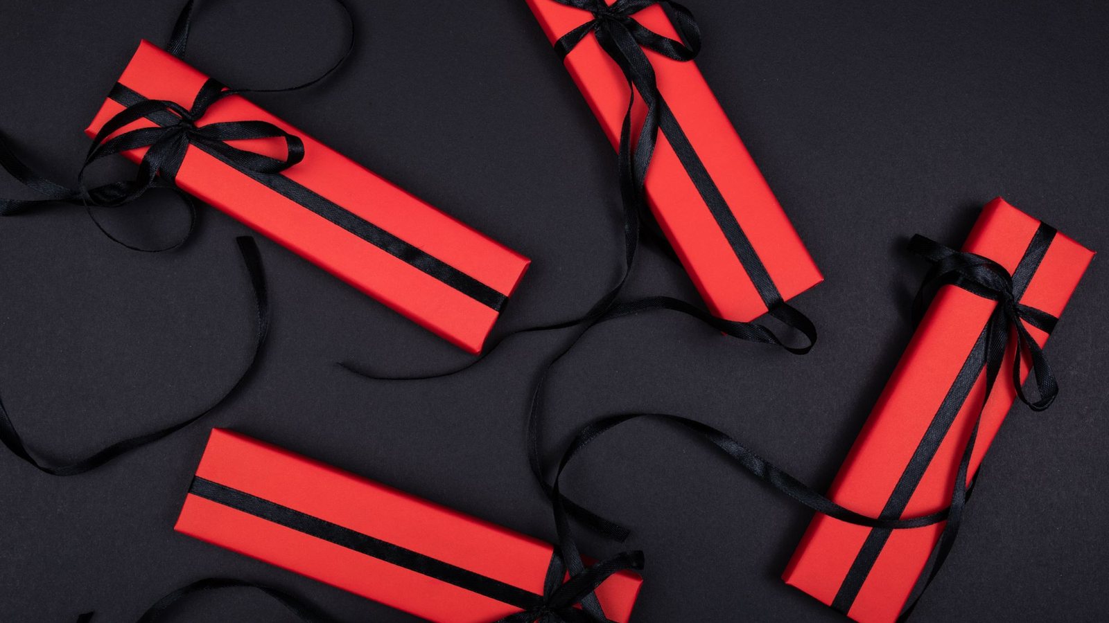 Photograph of slim red gift boxes tied with black ribbon scattered on a black surface