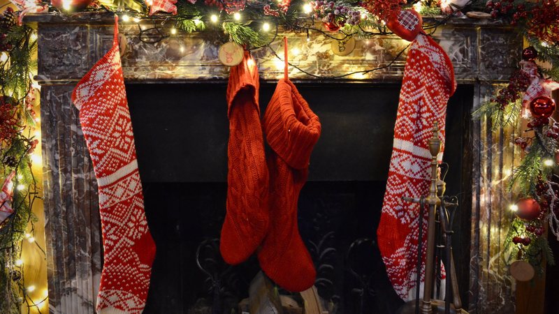 Photograph of red and white knit stockings hanging from a marble fireplace mantel with a lit garland