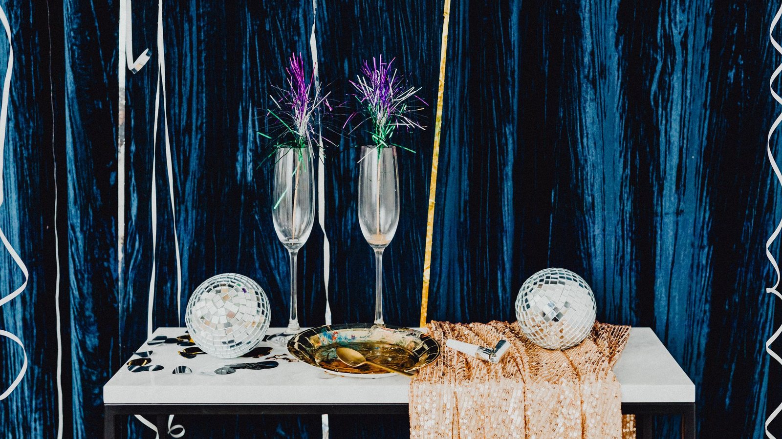 Photograph of two Champagne flutes, mirror balls and a gold tray on a white table in front of a dark blue velvet curtain