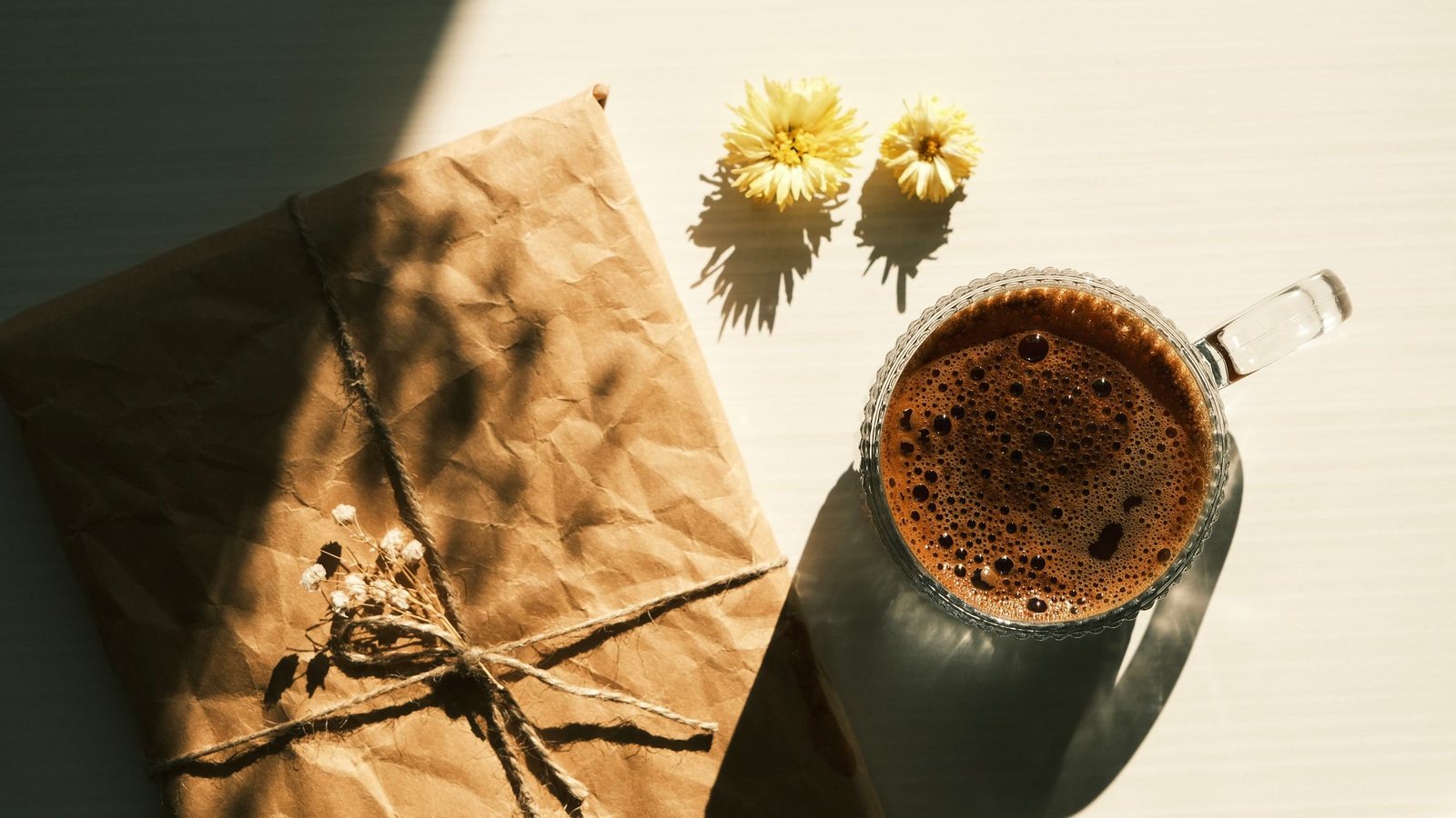 Photograph of a parcel wrapped in brown paper and twine beside a glass of coffee and two small yellow flowers in bright sunlight