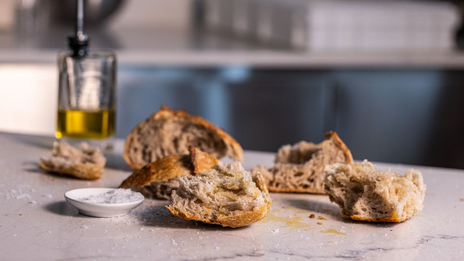 Photograph of torn rustic bread, a dish of salt and a glass cruet of olive oil on a marble kitchen counter